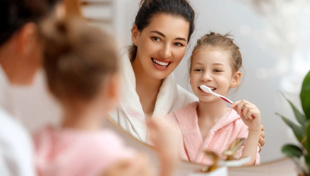 Family Brushing Teeth Together – Preventive Dental Care – Tesoro Dental Mother and daughter smiling while brushing their teeth together, highlighting the importance of family oral hygiene and preventive dental care for children and adults - Tesoro Dental
